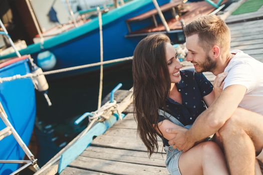 Lovers, guy and girl, are sitting on a wooden pier, holding hands and laughing.