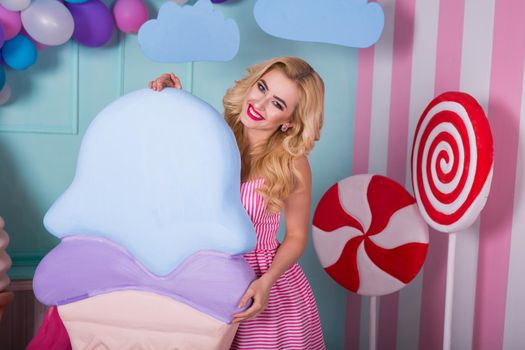 Portrait of young woman in pink dress holding big ice cream and posing on decorated background. Amazing sweet-tooth girl surrounded by toy sweets.