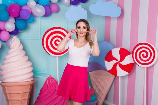 Portrait of joyful young woman in pink dress on background decorated with huge candies and ice cream.