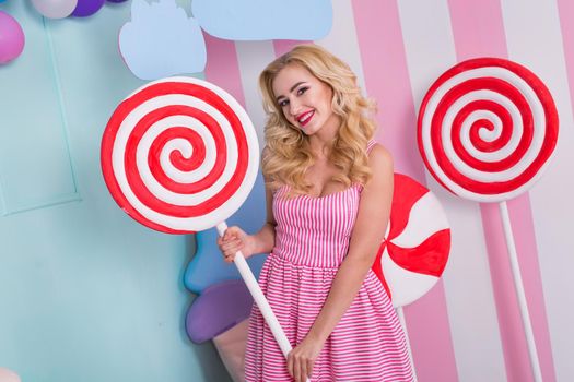 Portrait of young woman in pink dress holding big candy cane and posing on decorated background.