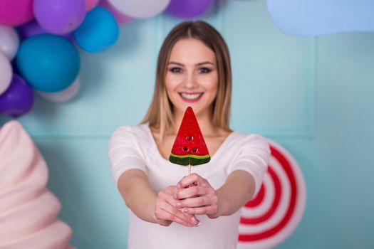 Portrait of amazing sweet-tooth woman in pink dress holding candies and posing on background decorated with huge ice cream. Lollipop watermelon