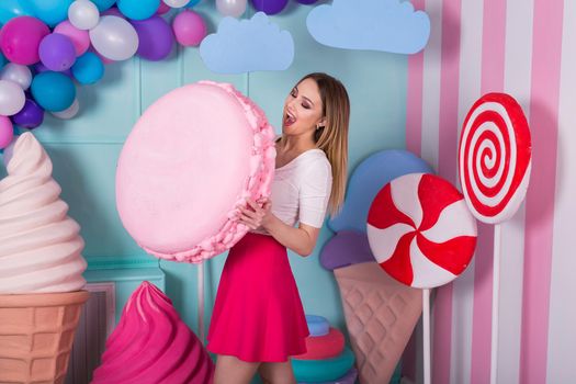 Portrait of young woman in pink dress holding big macaroon and posing on decorated background. Amazing sweet-tooth girl surrounded by toy sweets.