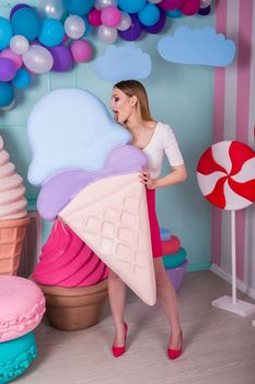 Portrait of young woman in pink dress holding big ice cream and posing on decorated background. Amazing sweet-tooth girl surrounded by toy sweets.
