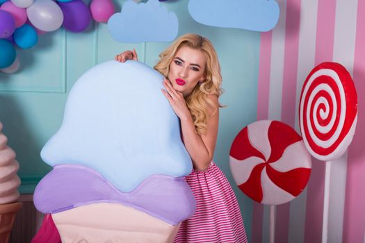 Portrait of young woman in pink dress holding big ice cream and posing on decorated background. Amazing sweet-tooth girl surrounded by toy sweets.