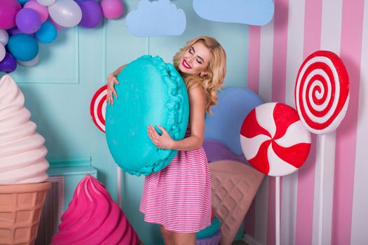 Portrait of young woman in pink dress holding big macaroon and posing on decorated background. Amazing sweet-tooth girl surrounded by toy sweets.