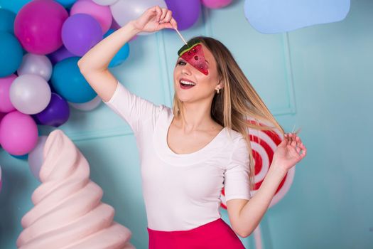 Portrait of amazing sweet-tooth woman in pink dress holding candies and posing on background decorated with huge ice cream. Lollipop watermelon