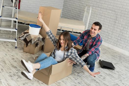 Happy couple having fun laughing moving into new home, young excited woman riding sitting in cardboard box while man pushing it, cheerful roommates playing while packing unpacking belongings together
