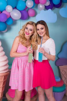 Two young women in pink dresses drink juice in a glass bottle.