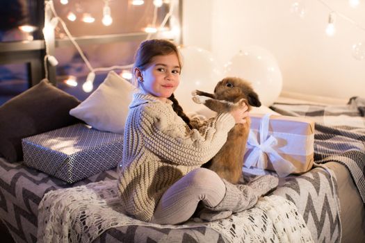 Very beautiful and charming little dark-haired girl in white sweater holds a live rabbit in the interior of the house. New year. Christmas. Hare. Studio.