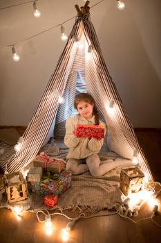Beautiful little girl sitting with a present in his hands. Deploy Christmas gift box on holiday in the evening in the beautiful interior room. New year. Studio