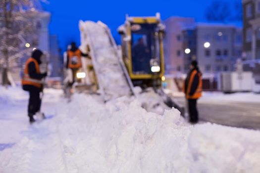 Claw loader vehicle removes snow from the road. Uniformed worker helps shovel snow into a snowplow.