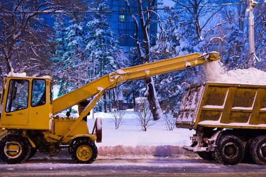 Claw loader vehicle removes snow from the road. A snowplow pours snow into a truck.