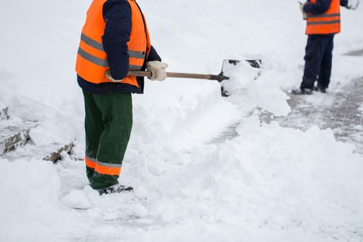 Employees of municipal services in a special form are clearing snow from the sidewalk with a shovel.