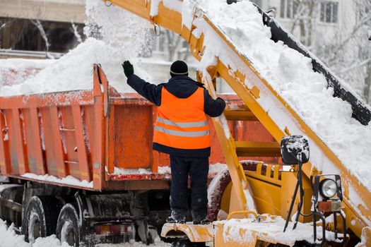 Claw loader vehicle removes snow from the road. Uniformed community service worker helps load snow into a truck.