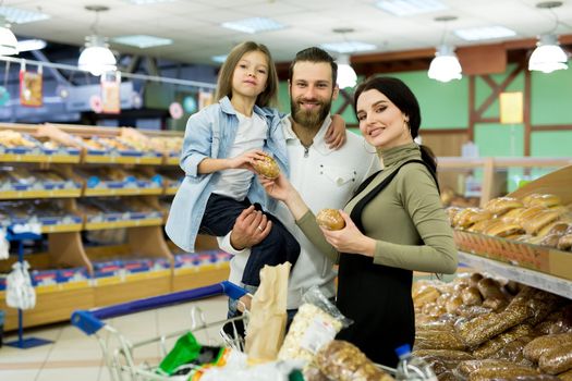 Beautiful young parents and their cute little daughter are smiling while choosing baking in the supermarket