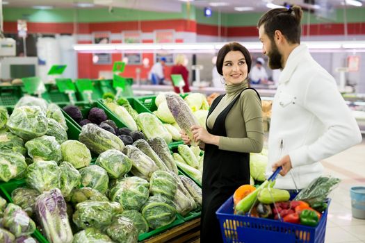 A young family, a man and a woman choose vegetables, cabbage in a large supermarket.