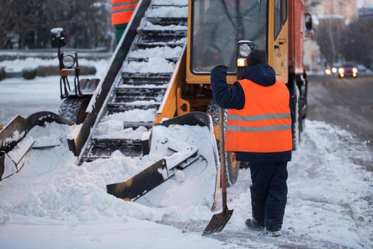 Tractor cleaning the road from the snow. Excavator cleans the streets of large amounts of snow in city. Workers sweep snow from road in winter, Cleaning road from snow storm.