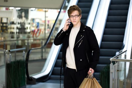 A young man in glasses with paper bags shopping at the Mall and talking on the phone.