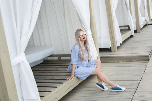 Portrait of pretty blonde girl with long hair posing to the camera on the street. She wears blue dress and glasses