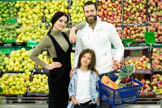 Portrait of a cheerful family standing with a full cart in the supermarket in the vegetable Department on the background of the counter with apples.