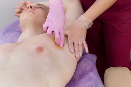 Close-up of the girl master sugaring causes a thick sugar paste on the armpits of a young sexy man. Male bodybuilder lying on the couch in the Spa salon for hair removal procedure