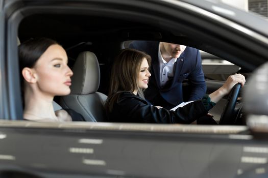 Gorgeous young girls sitting inside car, smiling and looking at camera. One of girls holding hands on steering wheel. Female customers of car dealership choosing auto in showroom.