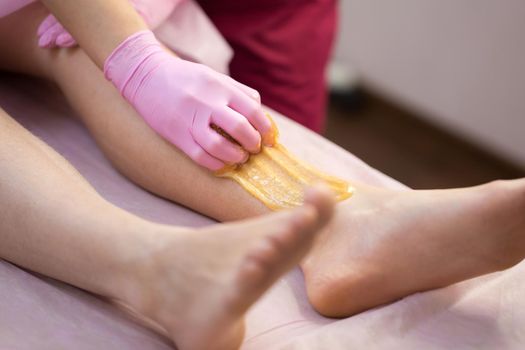 Sugar depilation of the feet in the beauty salon. Rid of hair on the legs. Sugaring. Master shugaring applies thick sugar paste on the legs of a young girl, removing hair on the legs. Close-up.