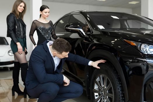 Attractive elegant man examining wheels of a new automobile on sale at dealership. Handsome male driver choosing new car to buy