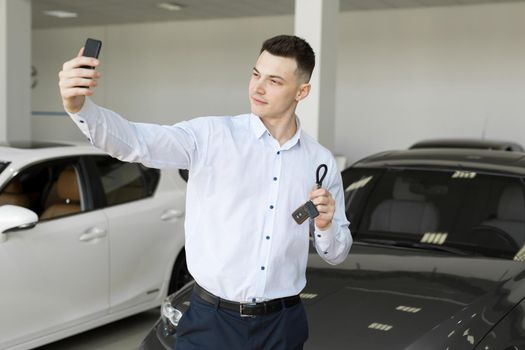 Happy businessman making selfie photo holding keys in front of his new car in the showroom