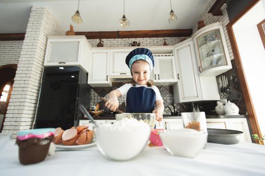 A cute little girl in an apron and a Chef's hat is stirring the dough with a wooden spatula, looking at the camera and smiling while cooking.