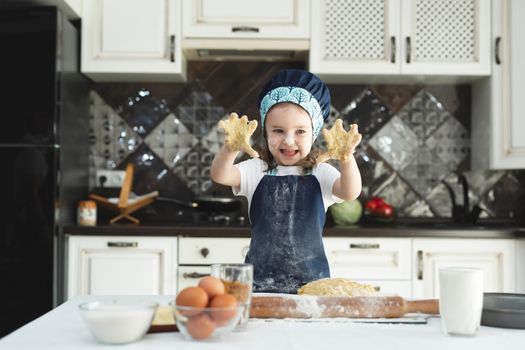 Little cheerful girl kneads dough with her hands in the kitchen in an apron and a chef's cap.