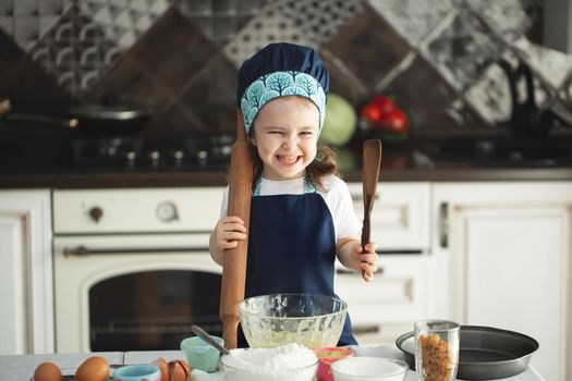 Cute little girl in apron and chef hat is flattening the dough using a rolling pin, looking at camera and smiling while baking