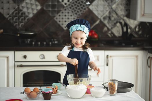 A cute little girl in an apron and a Chef's hat is stirring the dough with a wooden spatula, looking at the camera and smiling while cooking.