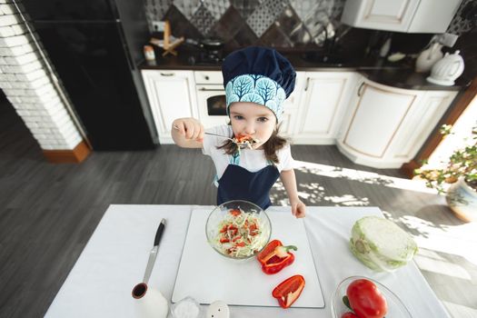 Child in an apron and a chef's hat is eating a vegetable salad in the kitchen and looking at the camera.
