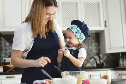 Cute little girl and her beautiful mom in matching aprons and caps play and laugh while kneading dough in the kitchen.