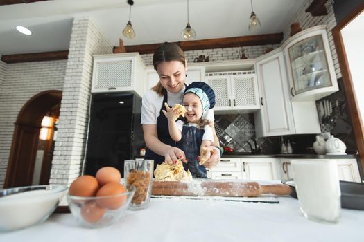 Cute small child daughter helping mum kneading preparing dough in bowl together in modern kitchen.