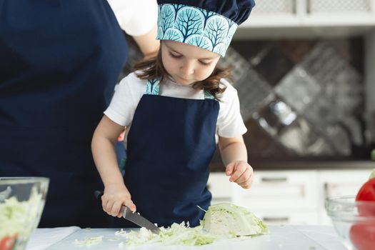 Caring mother teaching little daughter to cook salad in kitchen, young mum and adorable cute girl child wearing apron chopping vegetables with knife on countertop, standing in kitchen at home