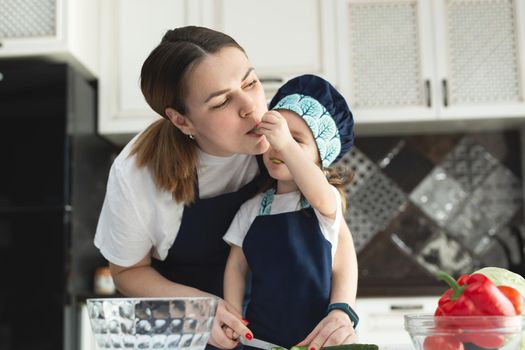 Caring mother teaching little daughter to cook salad in kitchen, young mum and adorable cute girl child wearing apron chopping vegetables with knife on countertop, standing in kitchen at home