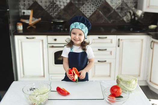 A child in an apron and a chef's hat in the kitchen is preparing a salad, eating red pepper and laughing.