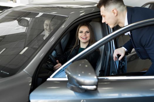 Side view of young beautiful woman sitting inside car and holding hand on steering wheel. She smiling and talking with manager of car dealership. Car agent representing inside of automobile.