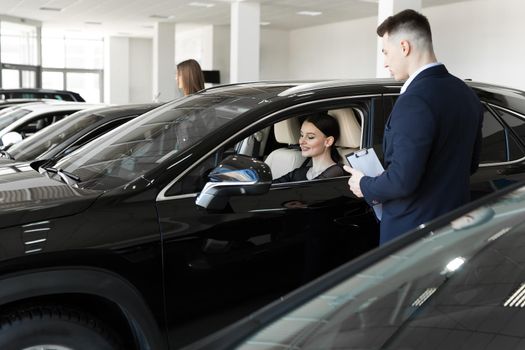 Side view of young beautiful woman sitting inside car and holding hand on steering wheel. She smiling and talking with manager of car dealership. Car agent representing inside of automobile.