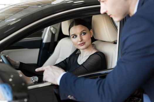 Side view of young beautiful woman sitting inside car and holding hand on steering wheel. She smiling and talking with manager of car dealership. Car agent representing inside of automobile.