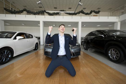 Young man in a suit is on his knees in a car dealership and is happy to buy a new car.