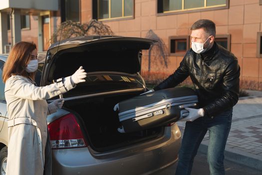 A man and a woman in protective medical masks and gloves load a suitcase into the trunk and leave the house by car during the quarantine and self-isolation. Coronavirus. covid 19.