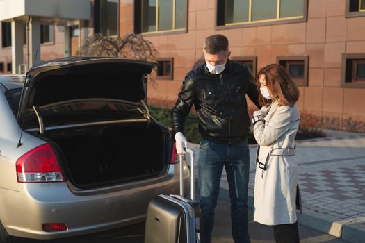 Man and woman in protective medical masks and gloves with a suitcase leave the house by car during the quarantine and self-isolation. The coronavirus. Covid 19.