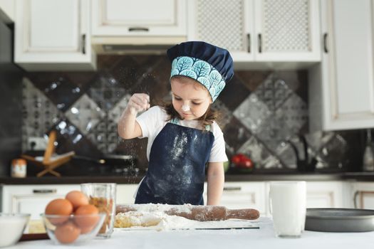 A little girl in an apron and a chef's hat sprinkle the dough with flour in the kitchen.