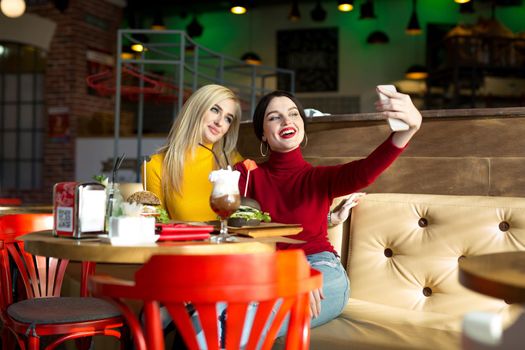 Two joyful cheerful girls taking a selfie while sitting together at cafe