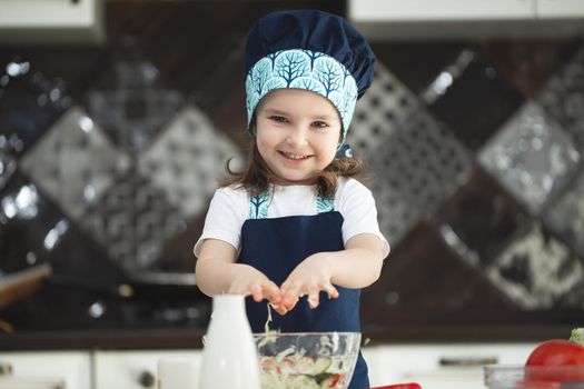 A child in an apron and a Chef's hat is stirring a vegetable salad in the kitchen and looking at the camera.