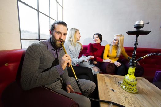 Friends smoke hookah and have fun, laugh. The man smiles and looks at the camera.