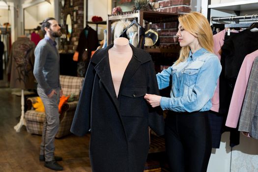 Beautiful young woman chooses a black coat in a clothing store. A man with a beard looks at her.